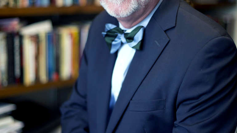 A white man with facial hair and hair pulled back staring ahead. He is in a blue suit with a blue and green bowtie. He is standing in front of a case of books.